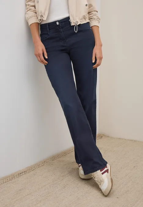 Frau in dunkelblauer Hose, beiger Jacke und Turnschuhen lehnt an einer Wand.